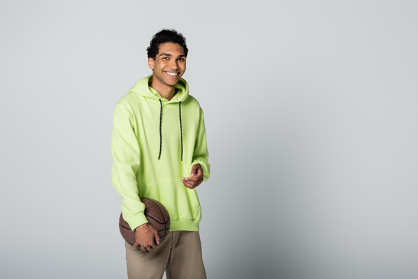 elegante hombre afroamericano con capucha verde sonriendo a la cámara mientras está de pie con baloncesto sobre fondo gris - Foto, Imagen