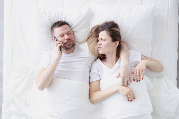 Young man lying in bed with his wife and talking on cell phone top view - Foto, Bild