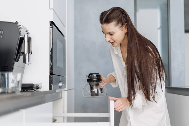 Young woman with long brunette hair in homewear taking tea pot from cupboard, preparing hot morning drink while standing in stylish fully furnished kitchen at home - Zdjęcie, obraz