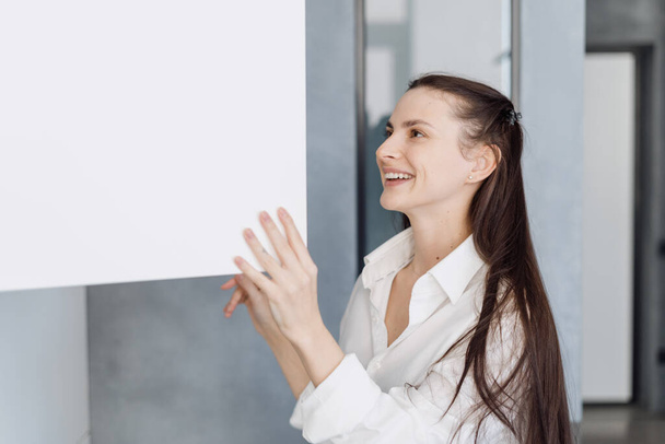 Счастливая молодая женщина в домашней одежде улыбается, открывая белый шкаф с чистой посудой и стоя утром на своей современной стильной кухне дома - Фото, изображение