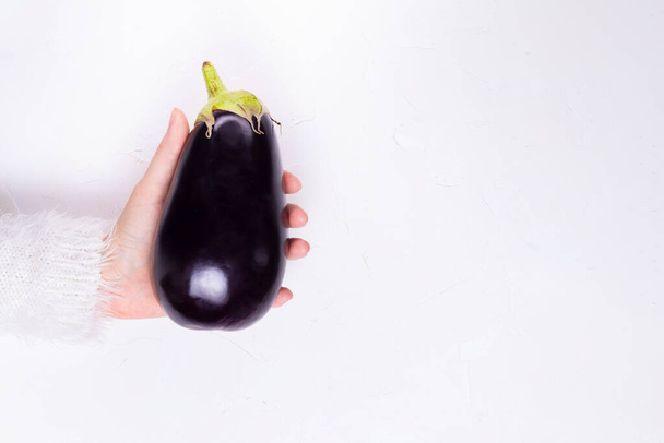 The hand of a young girl in a white fluffy sweater holds a ripe whole eggplant on a white background copy space - Fotó, kép