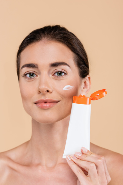 brunette woman with tube of sunscreen looking at camera isolated on beige - Foto, afbeelding
