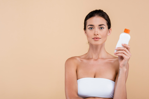 brunette woman with bottle of sunscreen looking at camera isolated on beige - Foto, afbeelding