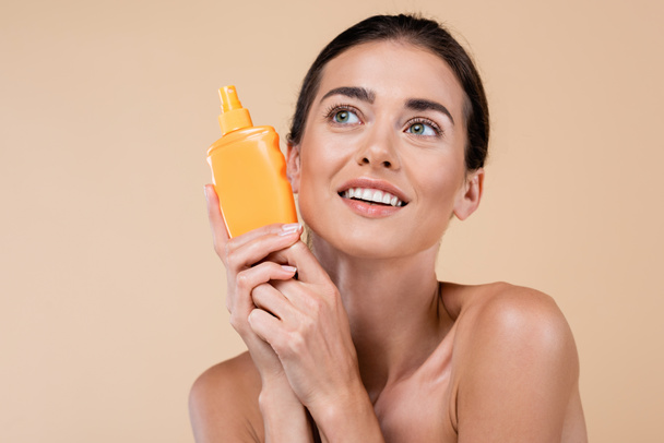 glimlachende vrouw met oranje fles zonnebrand weg te kijken geïsoleerd op beige - Foto, afbeelding