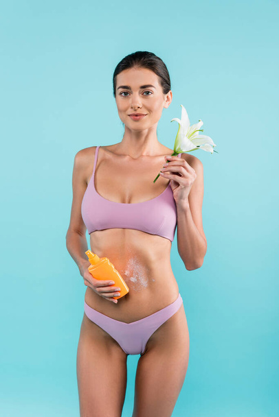slim woman with lily flower and sunblock looking at camera isolated on blue - Foto, Imagem