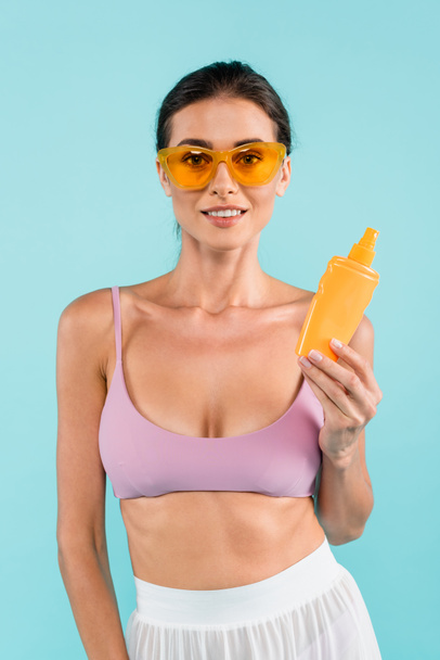 pleased woman in swimsuit and orange sunglasses holding sunblock isolated on blue - Foto, Bild