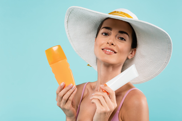 Güneş şapkalı neşeli kadın elinde tüp ve mavi üzerine izole edilmiş güneş kremi şişesiyle poz veriyor. - Fotoğraf, Görsel