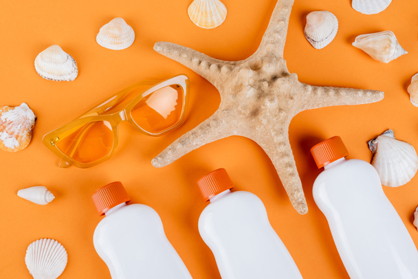 bovenaanzicht van zeesterren, schelpen, zonnebrillen en witte flessen zonnebrandcrème op oranje - Foto, afbeelding