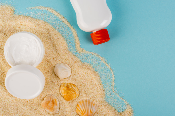 Sonnencreme und kosmetische Creme in der Nähe von Muscheln auf Sand und blauer Oberfläche - Foto, Bild