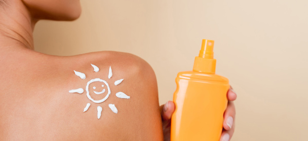 partial view of woman with bottle of sunblock and image of happy sun on naked shoulder isolated on beige, banner - Photo, Image