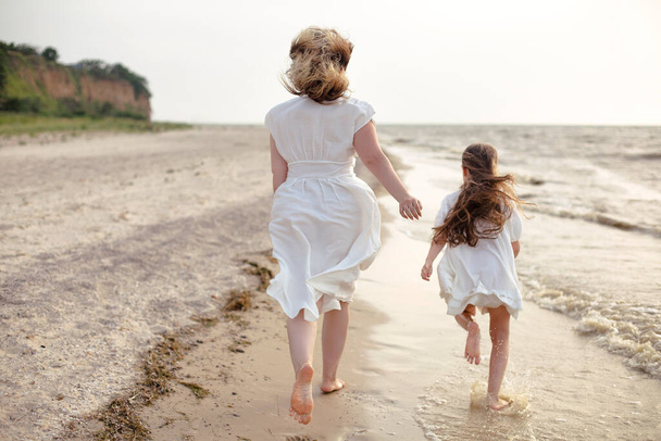Sabahları dalgalı denizin kenarında koşan beyaz elbiseli küçük kızı olan neşeli bir kadın. - Fotoğraf, Görsel