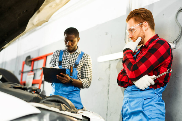 African American and red-haired craftsmen stand in overalls over the open hood of the car, defining the problem and thinking - Photo, image