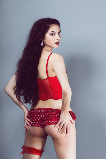 Sexy chica morena mira hacia atrás mientras está de pie y sosteniendo su culo en bragas rojas y tacones de corsé con liga en el estudio
 - Foto, imagen