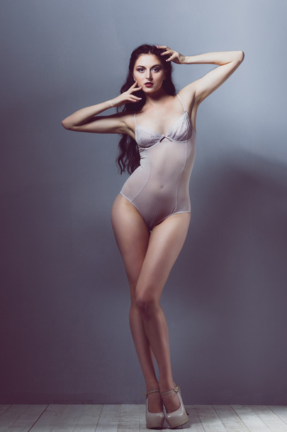 το σέξι κορίτσι μελαχρινή σε ένα διαφανές σώμα μπεζ τακούνια με ένα όμορφο σώμα, μακριά πόδια και μαλλιά ανάπτυξη με ένα άνεμο στο studio σε λευκό ξύλινο δάπεδο και γκρι τοίχους - Φωτογραφία, εικόνα