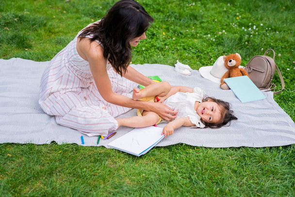 Vista laterale della giovane donna appoggiata in avanti al bambino stanco sdraiato su una coperta sull'erba verde mentre trascorreva la giornata estiva nel parco - Foto, immagini