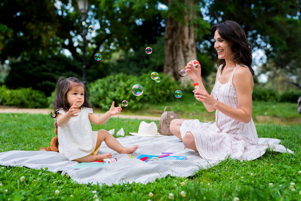 Cheerful young mommy blowing bubbles and watching curious little daughter playing while sitting on blanket together in park in summer - Photo, Image