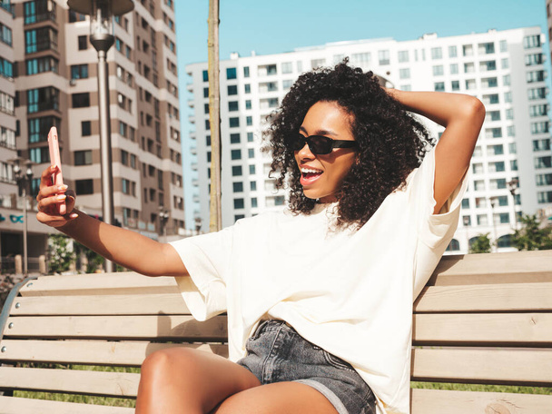 Beautiful black woman with afro curls hairstyle.Smiling hipster model in white t-shirt. Sexy carefree female posing in the street in sunglasses. Cheerful and happy.Taking selfie photo - 写真・画像