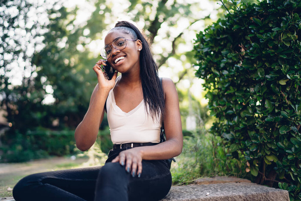 From below of happy young black female in casual clothes and glasses talking on mobile phone while sitting on stone step in summer park - Fotoğraf, Görsel