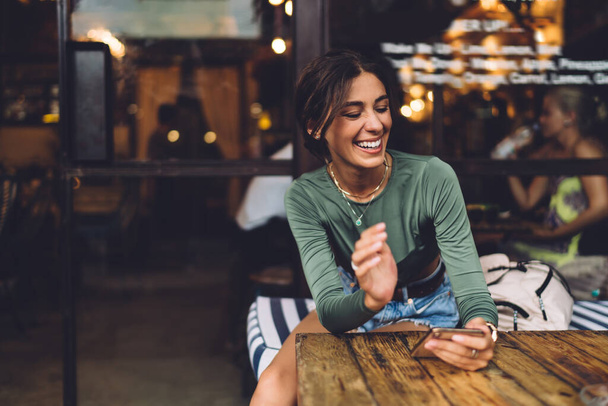 Optimistic female in casual outfit smiling and chatting on mobile phone while resting at wooden table in cozy cafe during weekend - Fotoğraf, Görsel