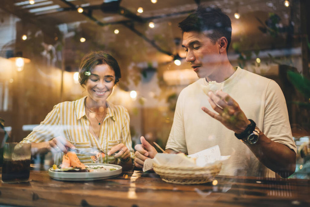 Through window of positive man and woman having friendly conversation in cozy restaurant while eating tasty food and drinking beverages - 写真・画像
