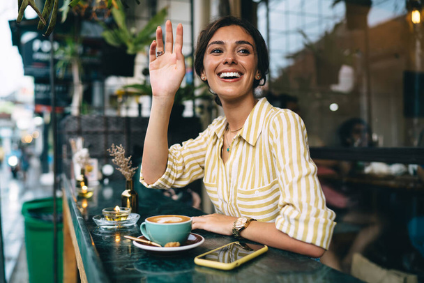 Happy ethnic female in trendy casual clothes smiling and raising hand for greeting while sitting at table with cup of coffee and smartphone in street cafe - Foto, Imagem