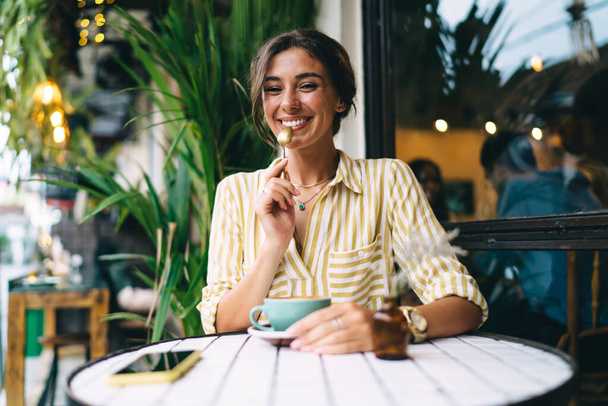 Optimistic young female wearing striped shirt drinking cup of fresh coffee and holding teaspoon while sitting at round table in open terrace - Foto, Bild