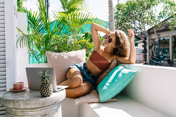 Content relaxed female in casual wear and sunglasses sitting on sofa on sunny terrace on Bali resort and enjoying summer - Foto, Bild