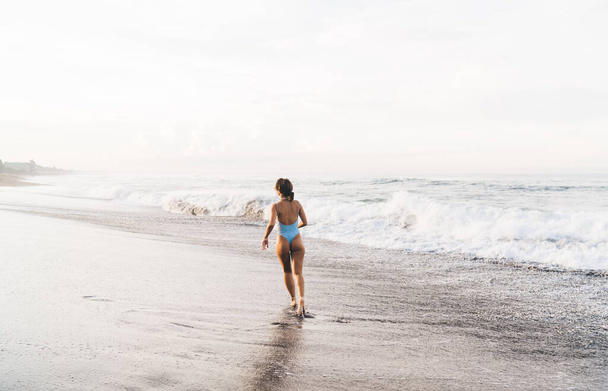 Back view of anonymous female in swimsuit walking on wet shore near waving sea and enjoying sunny summer day alone - Foto, Imagem