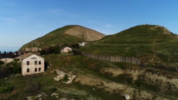 Aerial view of the abandoned village located between the hills. Shot. Picturesque mountains and cliffs covered by green meadow with rare old houses on blue sky background. - Footage, Video