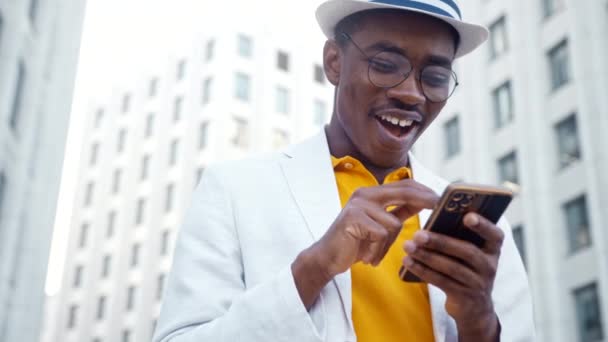 Hombre negro en traje con tipos de sombrero en el teléfono inteligente y se ríe - Imágenes, Vídeo