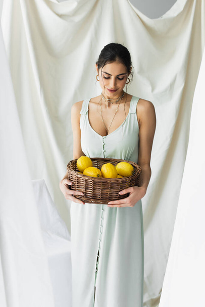 smiling woman in dress looking ripe lemons in wicker basket on white - Foto, afbeelding
