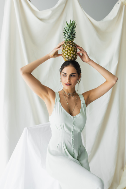 brunette woman in dress posing with pineapple above head on white  - Foto, afbeelding