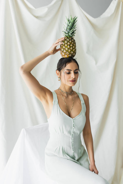 brunette woman in dress holding pineapple above head on white  - Foto, afbeelding