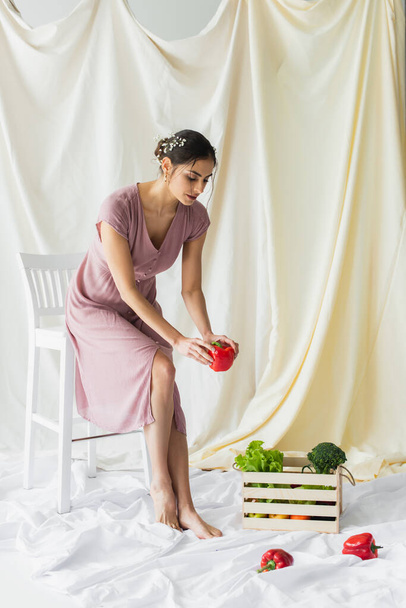 брюнетка жінка тримає червоний болгарський перець біля овочів у дерев'яній тарілці на білому
 - Фото, зображення