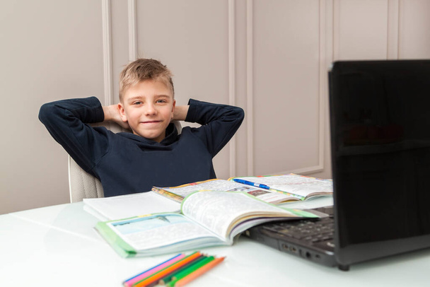 happy boy leans back in chair and puts hands behind head, sitting at table with notebooks, textbooks and pencils in front of laptop. Smart Little Boy doing His homework in Living Room. COVID-19 Pandemic forces Kids to Learn online - Zdjęcie, obraz