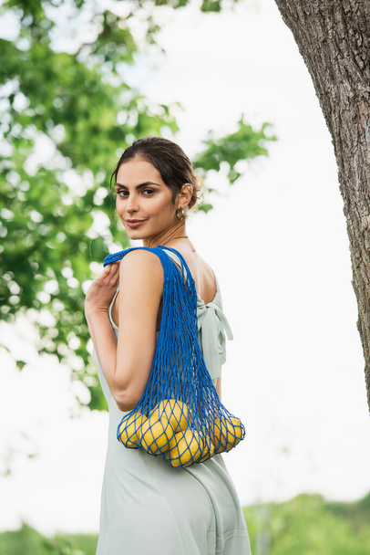 brunette woman holding reusable string bag with lemons in park - Photo, image