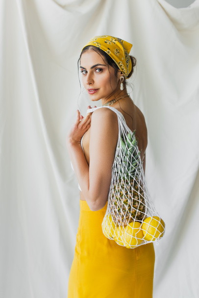 woman in yellow headscarf and crop top holding string bag with lemons and pineapple on white - Фото, зображення