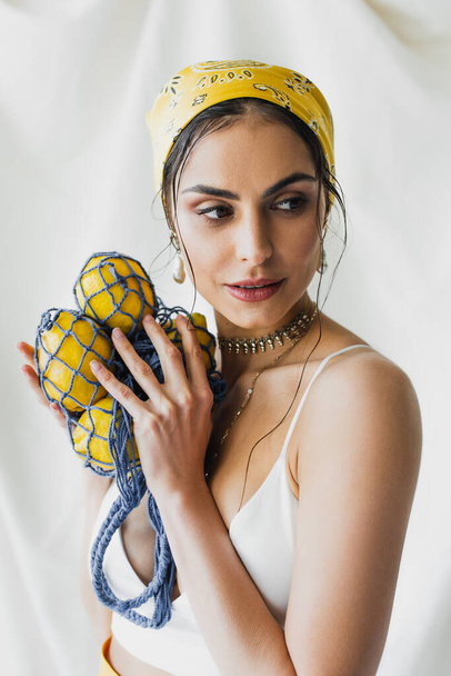 woman in yellow headscarf and crop top holding string bag with lemons on white - Fotoğraf, Görsel