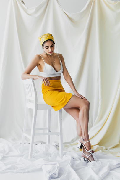 full length of stylish woman in yellow headscarf, skirt and crop top sitting on chair on white - Photo, Image