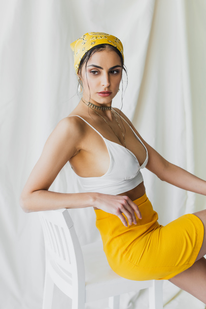 stylish woman in yellow headscarf, skirt and crop top sitting on chair on white - Photo, Image