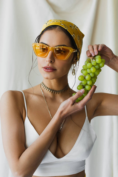 trendy woman in yellow sunglasses, headscarf and crop top holding grapes on white  - Foto, Bild