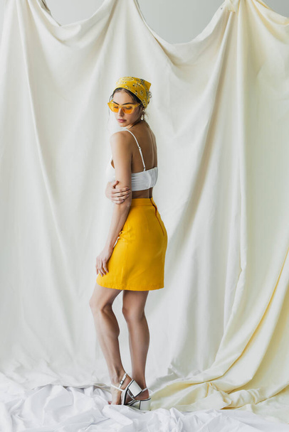 full length of pretty woman in yellow sunglasses and headscarf posing on white - Zdjęcie, obraz