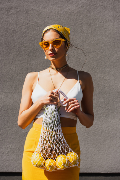 mujer con estilo en gafas de sol amarillas y pañuelo para la cabeza bolsa de cuerda con frutas frescas cerca de la pared de hormigón  - Foto, Imagen