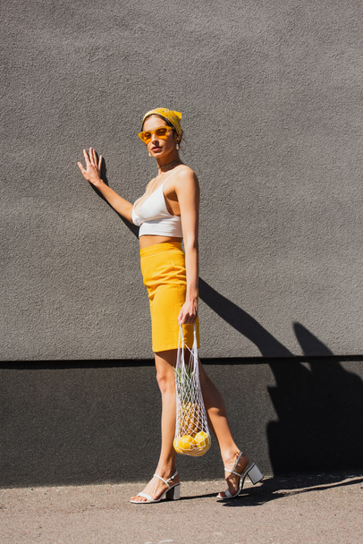 full length of trendy woman in sunglasses and yellow headscarf holding string bag with fruits near concrete wall  - Valokuva, kuva