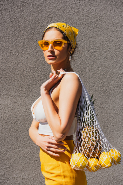Trendfrau mit Sonnenbrille und gelbem Kopftuch, die in der Nähe einer Betonmauer eine Tasche mit Früchten in der Hand hält  - Foto, Bild