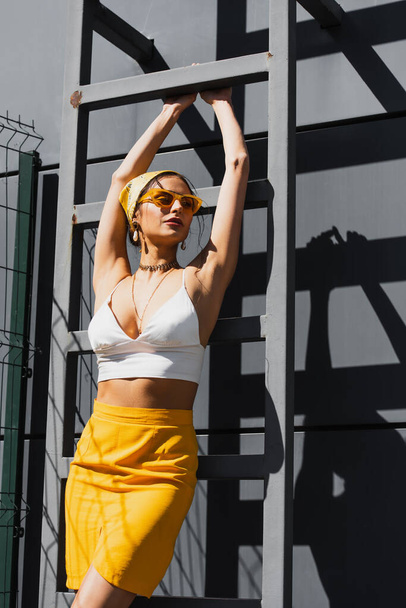 ładna kobieta w okularach przeciwsłonecznych i żółtej chustce na głowie, pozująca blisko drabiny i betonowej ściany  - Zdjęcie, obraz