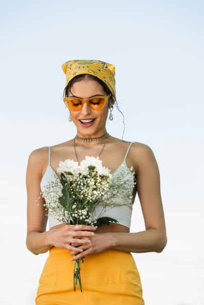 изумленная женщина в солнечных очках и желтом платке, смотрящая на цветы  - Фото, изображение