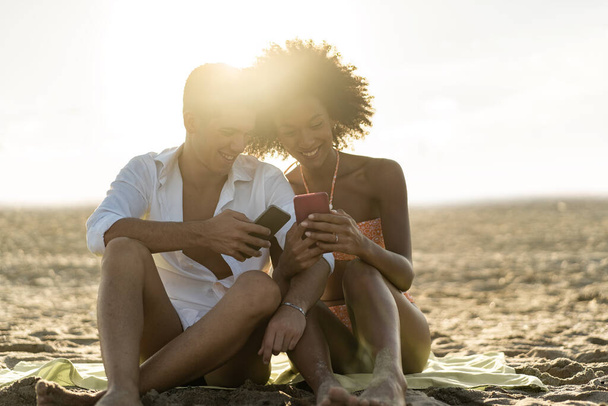 Interrassisches Paar, das Spaß daran hat, Social-Media-Inhalte auf dem Smartphone-Bildschirm am Strand bei Sonnenuntergang zu sehen - Foto, Bild