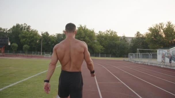 A muscular man with a naked torso walks along the treadmill of the city stadium. Back view. Slow motion - Footage, Video