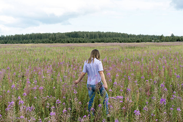 Mulher loira bonita nova na camisa roxa de trás andando no prado entre flores de fireweed, beleza na paisagem da natureza - Foto, Imagem